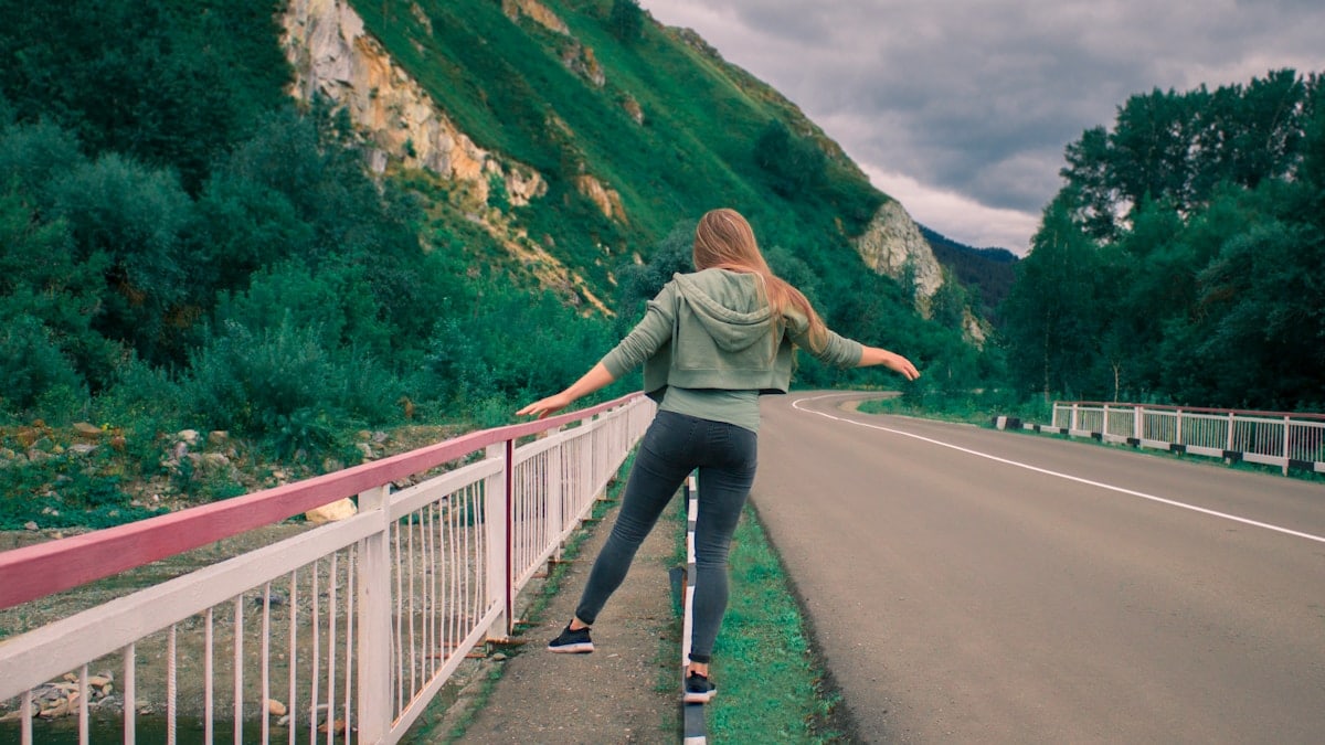 woman in gray jacket and blue denim jeans walking on gray concrete bridge during daytime